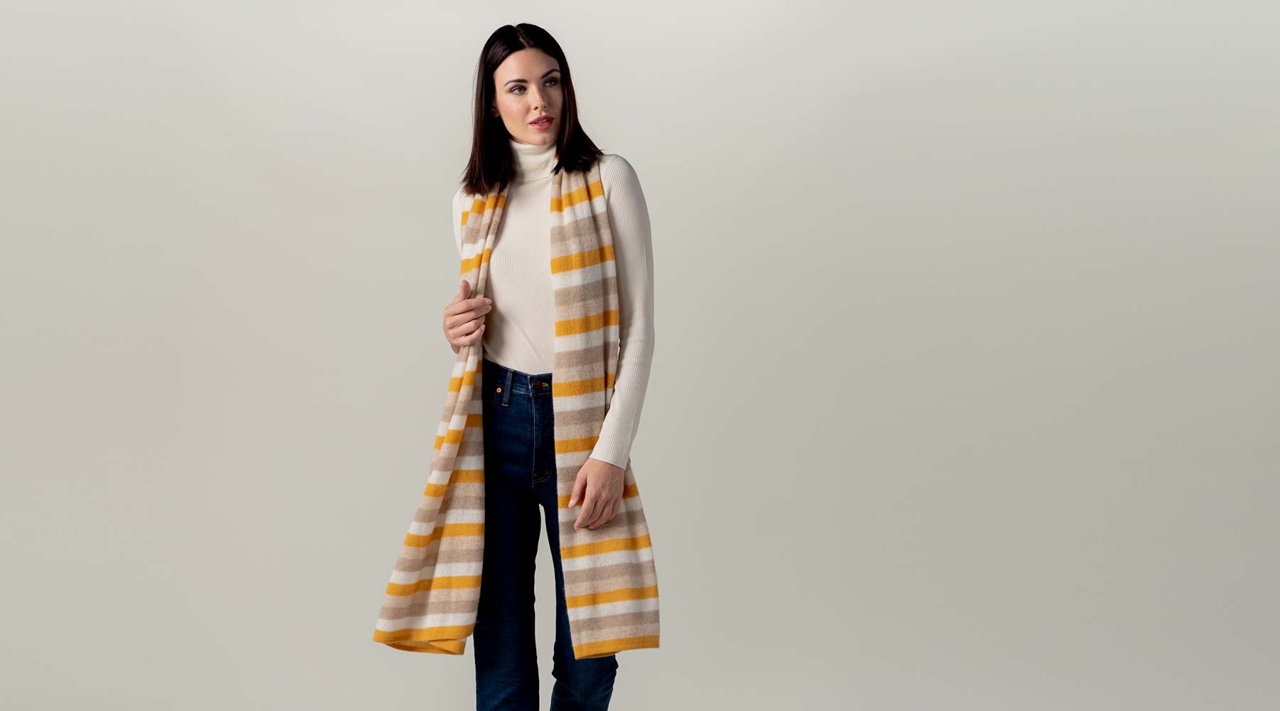 A dark-haired woman wearing a cream turtleneck and striped cashmere wrap scarf in yellow, beige, and cream tones.