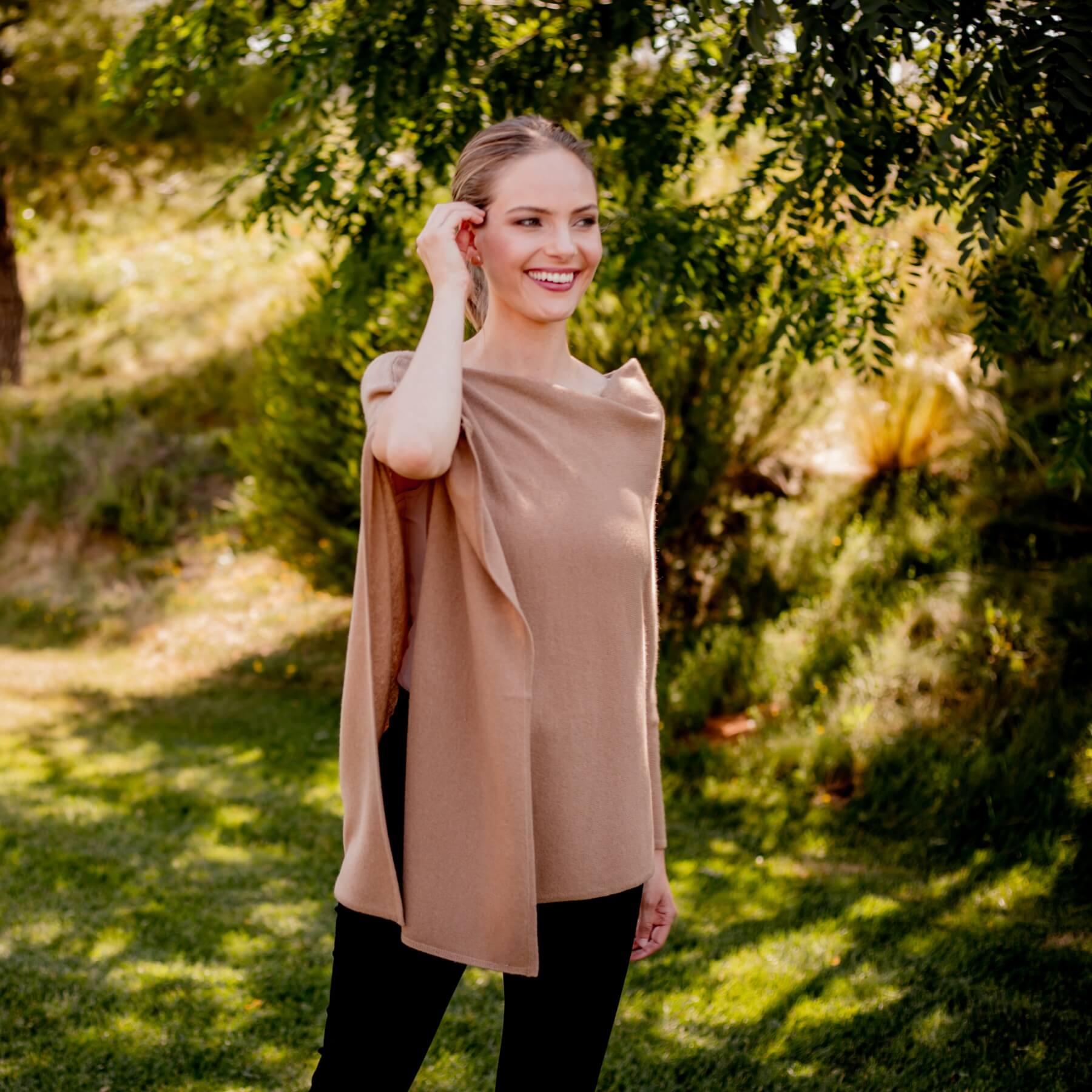 A woman wearing a draped, short-sleeved camel-colored top or poncho with black pants, standing outdoors in a grassy area with trees in the background.